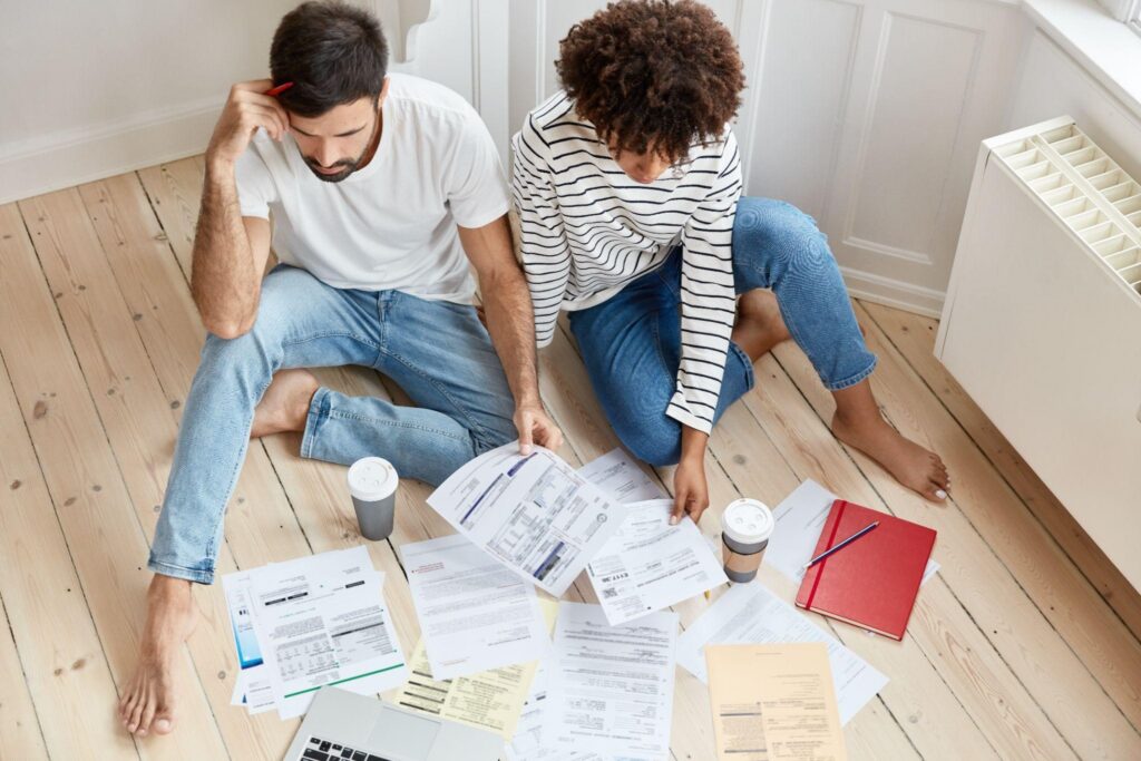 Two people sit on a wooden floor surrounded by scattered papers and notebooks, reviewing documents together.