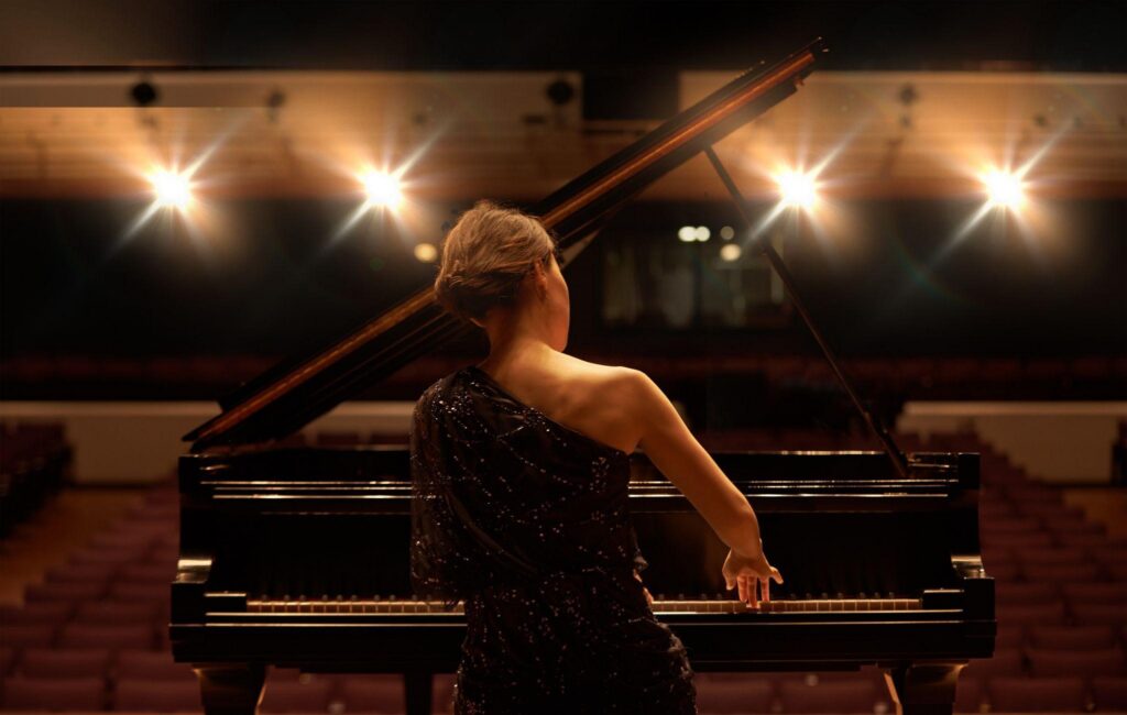 Back view of a pianist in a sequined gown playing a grand piano on a warmly lit stage.