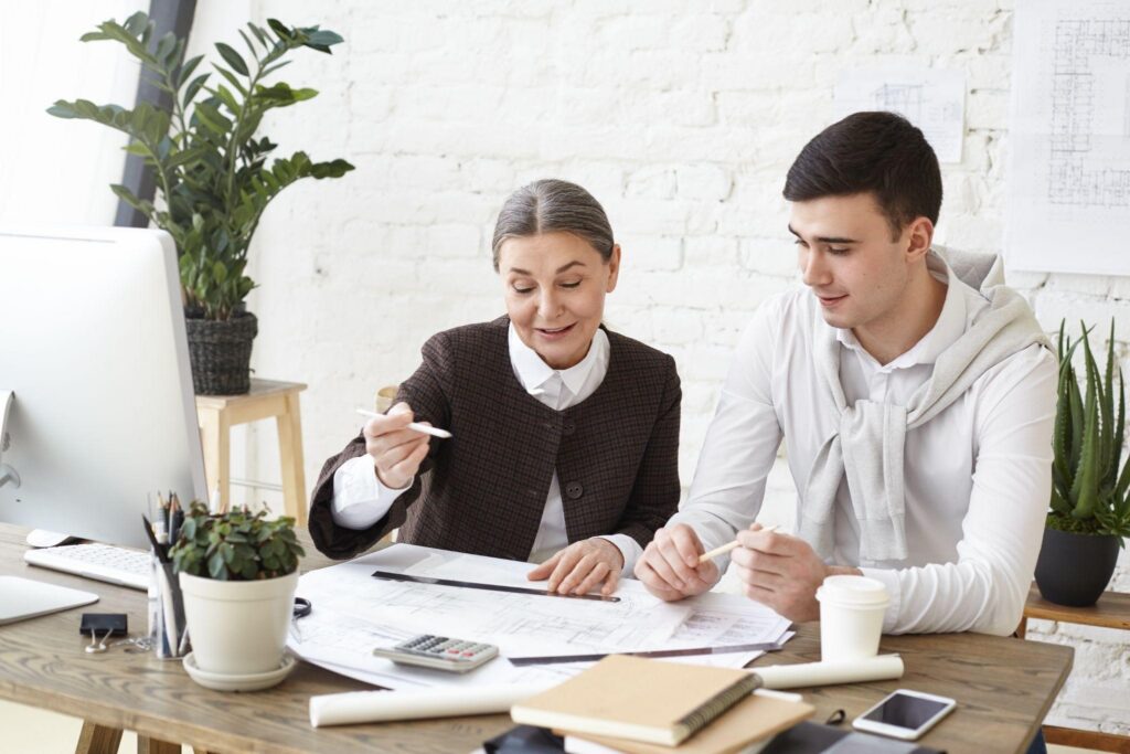 Two professionals review architectural blueprints at a wooden desk in a bright office, with a computer, plants, and coffee nearby.