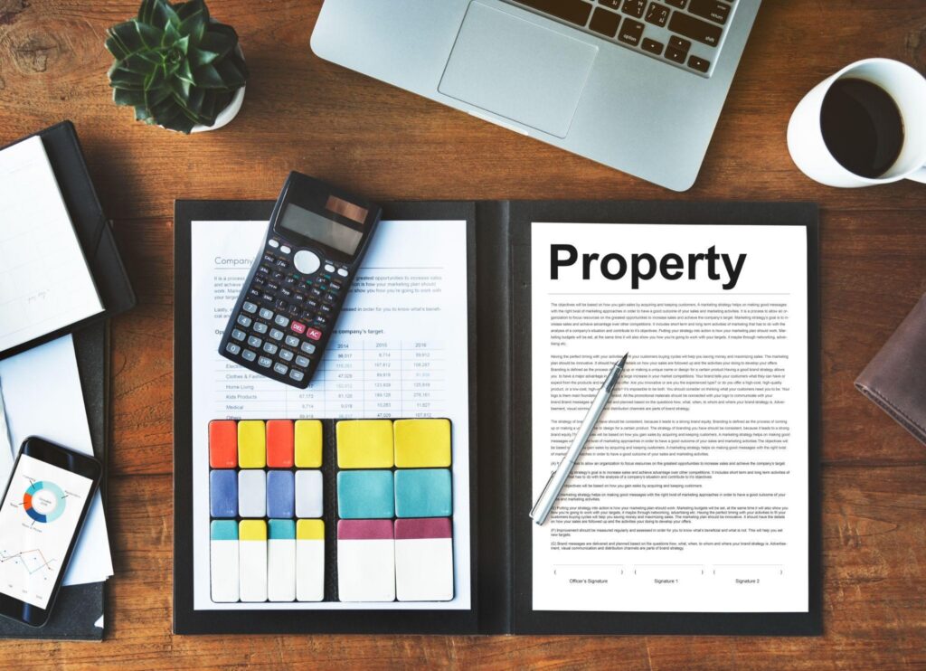A top-view office desk with a laptop, coffee cup, plant, calculator, and an open binder labeled 'Property' with colorful tabbed pages